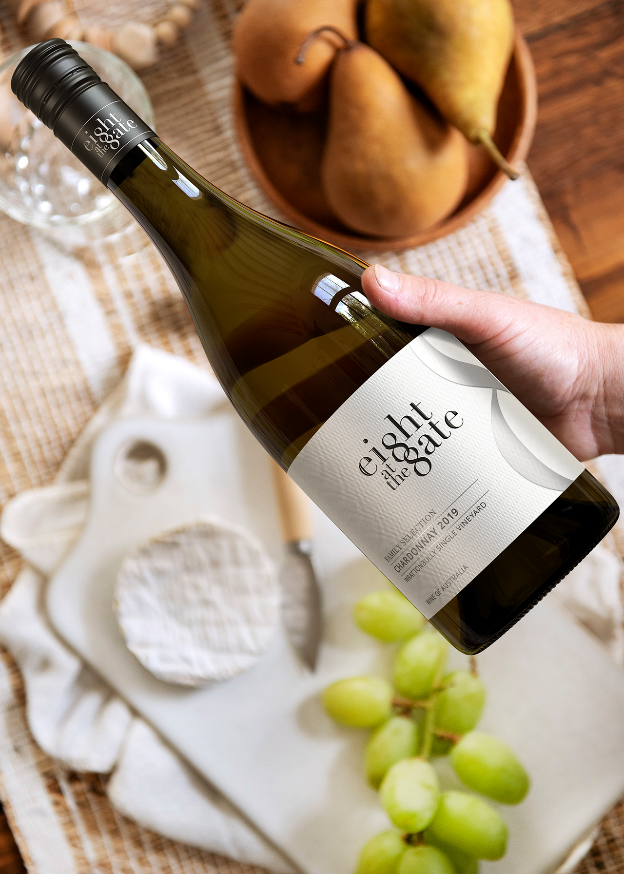 A hand holding a bottle of Eight at the Gate Family Selection Chardonnay Single Vineyard 2019 over a cheese board.